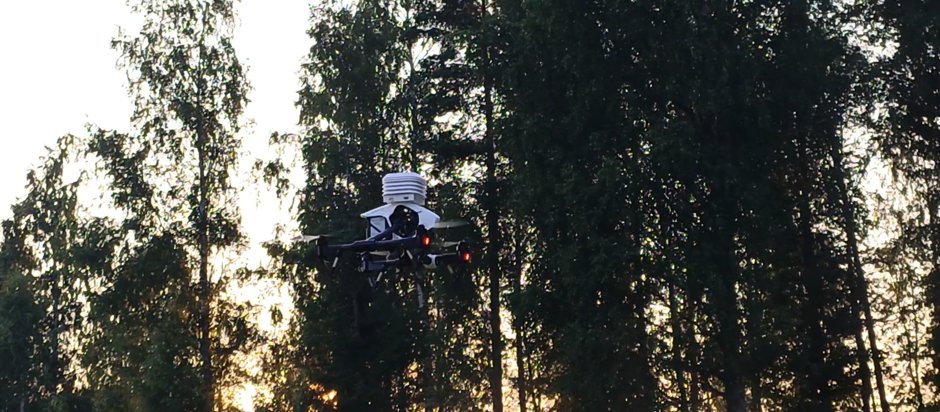 A WeatherSens AI drone hovering at treetop height against a low sun, its meteorological sensor shield on top.